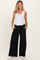 woman wearing a white tank top and black wide-leg maternity pants with pockets, standing with hands in pockets