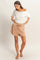 woman wearing a white off-shoulder top and beige drawstring shorts, standing barefoot and looking down