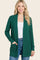 woman wearing a green long sleeve cardigan with pockets and a white top, smiling against a neutral background