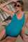 woman wearing a teal maternity swimsuit sitting on the beach and shading her eyes with sunglasses, baby bump visible