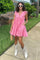woman wearing a pink ruffled sundress and white cowboy boots, holding a straw bag, standing on a sidewalk with greenery around