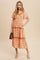 woman wearing a pink and orange floral print maxi dress with short sleeves and tiered ruffle details, holding a brown clutch