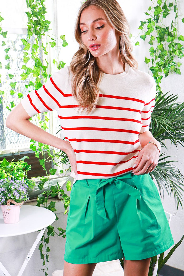 woman wearing a cream striped sweater with red stripes and green high-waisted shorts, posing in a vibrant indoor setting