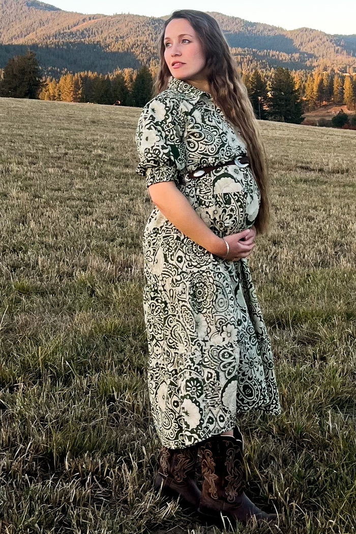 woman wearing a green and white paisley print maternity dress, standing in a field with a visible baby bump