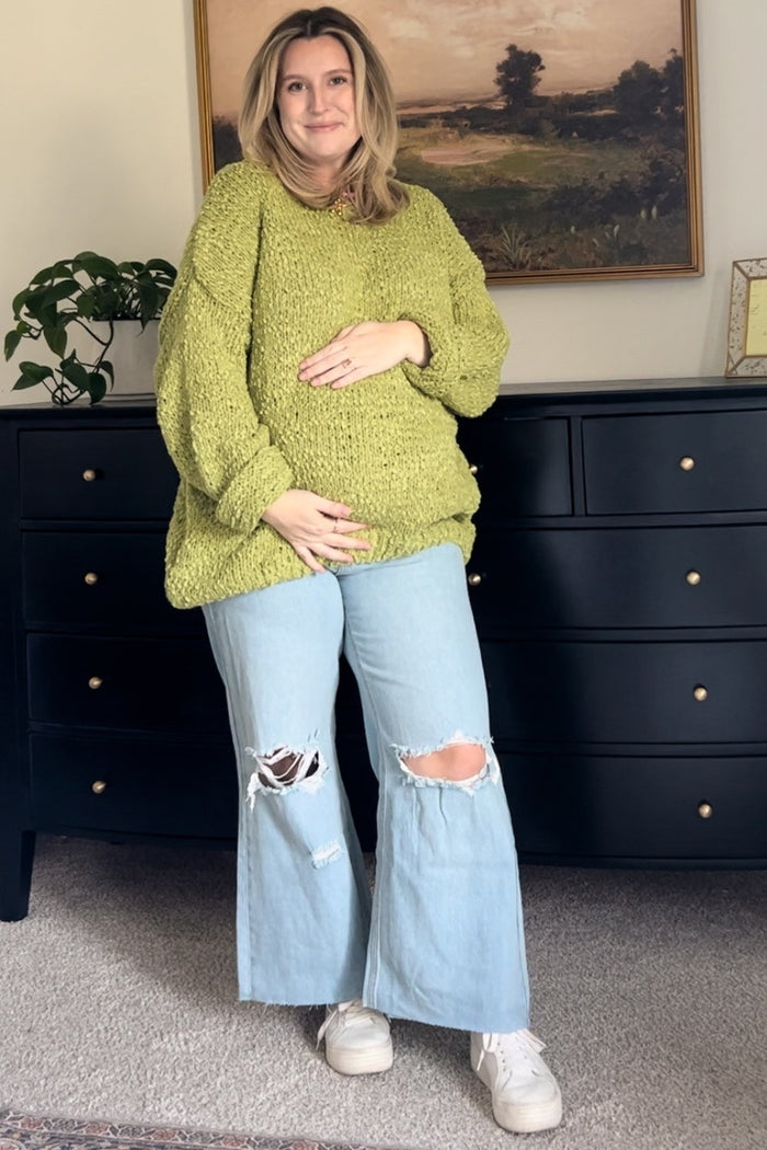 woman wearing a chunky green maternity sweater and distressed wide-leg jeans, cradling her baby bump