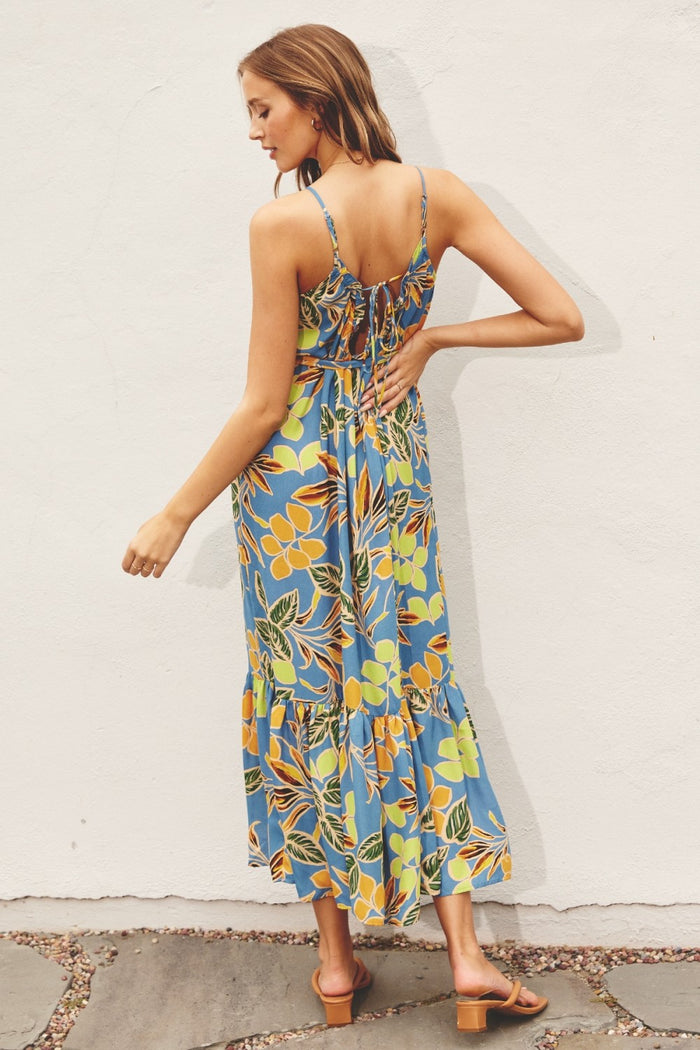 woman wearing a blue floral maxi dress with adjustable straps and a tiered hem, standing against a white wall