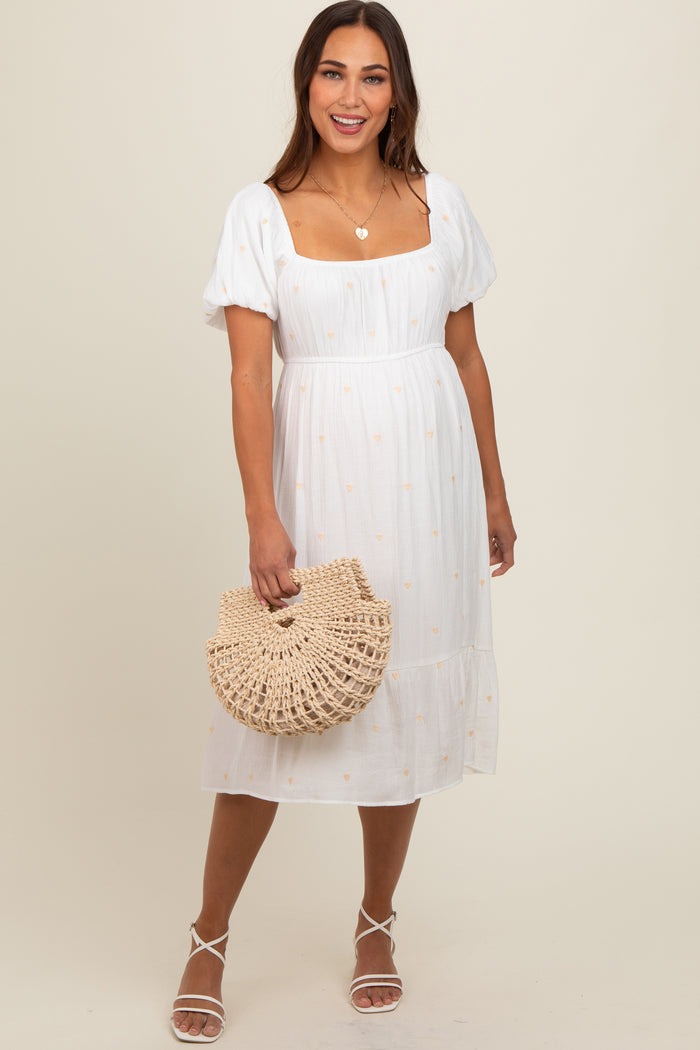 woman wearing a white maternity dress with puff sleeves and embroidered details, holding a woven bag