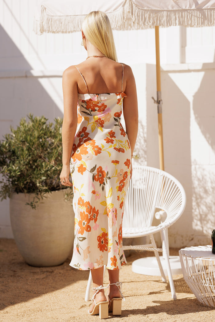 woman wearing a white floral midi dress with orange and pink flowers, seen from the back, standing outdoors by a plant