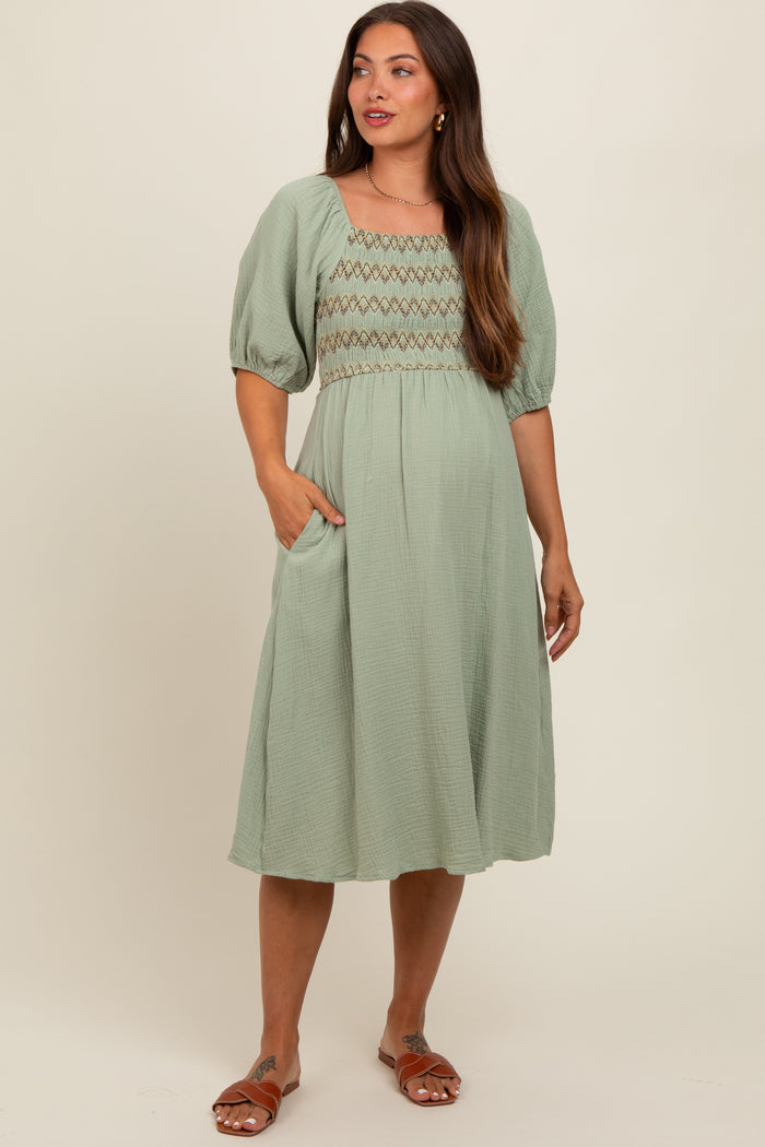 woman wearing a sage green maternity dress with puff sleeves and smocked bodice, posing with one hand in pocket