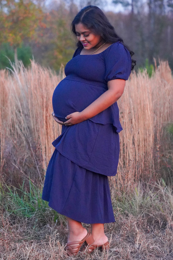 woman wearing a navy blue maternity two-piece outfit with a tiered skirt, holding her baby bump in a grassy field