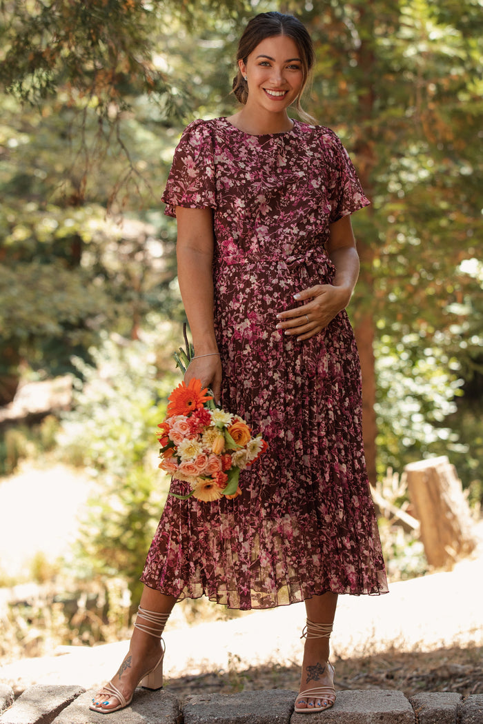 woman wearing a floral pleated maternity dress and holding a bouquet, with a visible baby bump