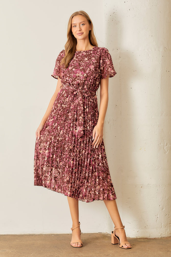 woman wearing a burgundy floral pleated midi dress with short sleeves and a tie waist, standing against a white wall