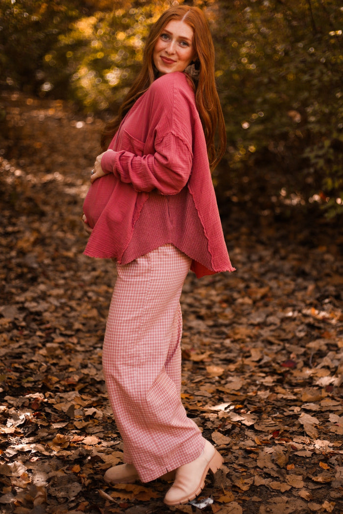 woman wearing a pink maternity top and checkered maternity pants, showing her baby bump in a forest setting