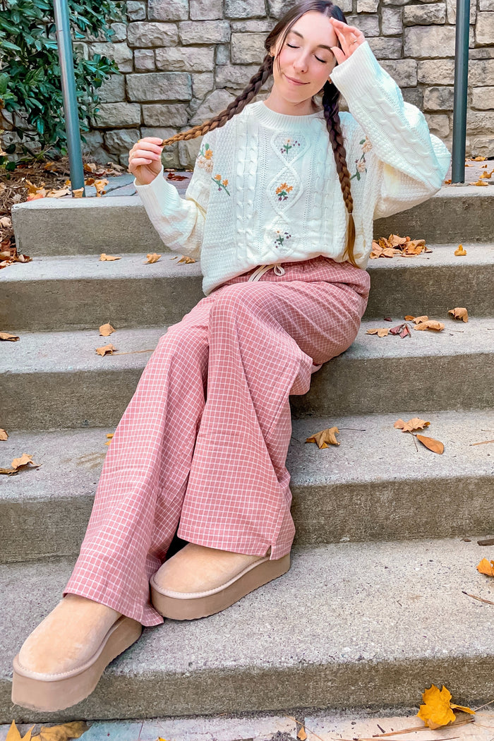woman wearing a cream cable knit sweater with floral details and pink checkered wide-leg pants, sitting on steps