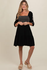 woman wearing a black maternity dress with embroidered details and a tiered skirt, smiling at the camera