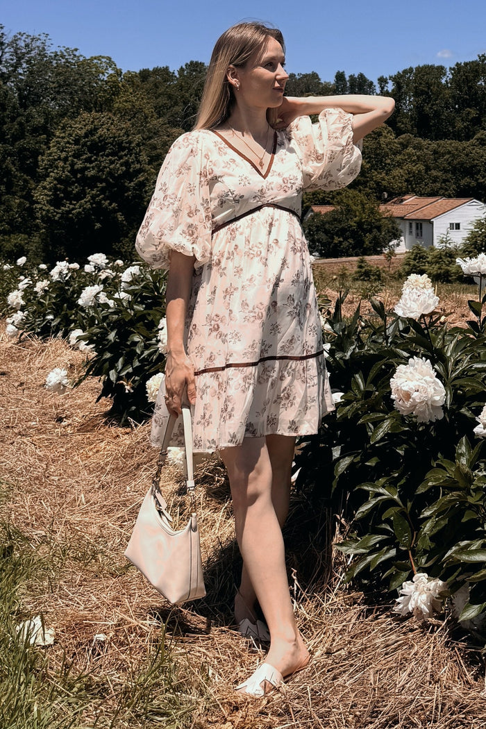 woman wearing a floral maternity dress with puff sleeves and a drawstring waist, holding a purse while standing in a flower field
