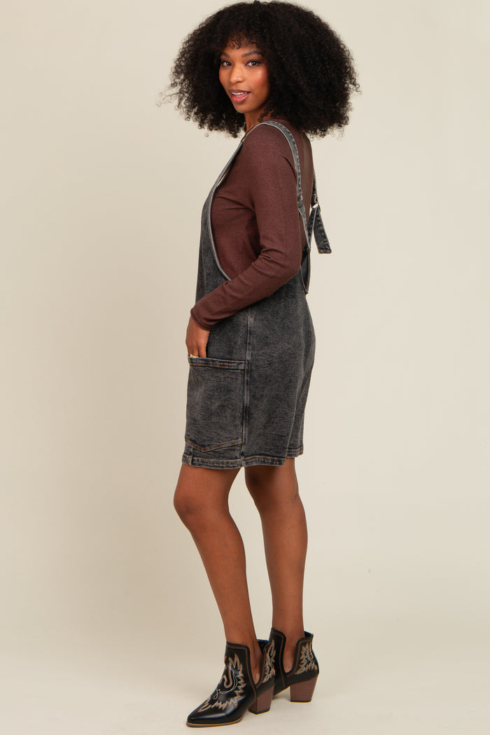 woman wearing a black denim overall dress over a brown long sleeve top, styled with ankle boots