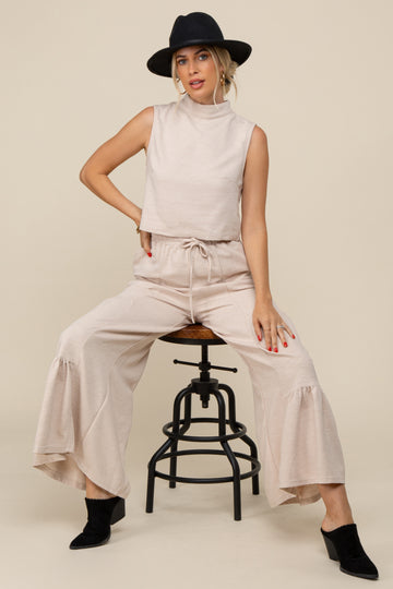 woman wearing a beige sleeveless mock neck top and matching wide-leg pants, sitting on a stool with a black hat and ankle boots