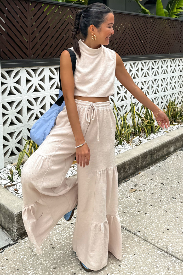 woman wearing a beige two-piece crop top and wide-leg pants set with ruffle hem details, smiling outdoors