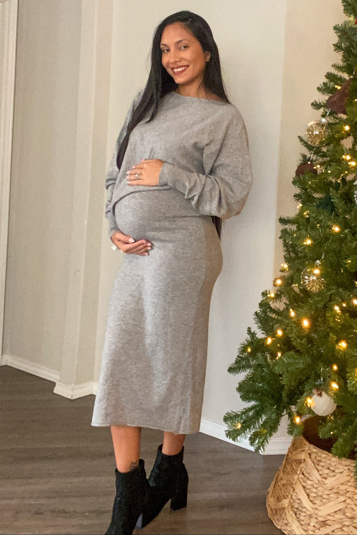 woman wearing a grey maternity dress and holding her baby bump, standing next to a decorated Christmas tree