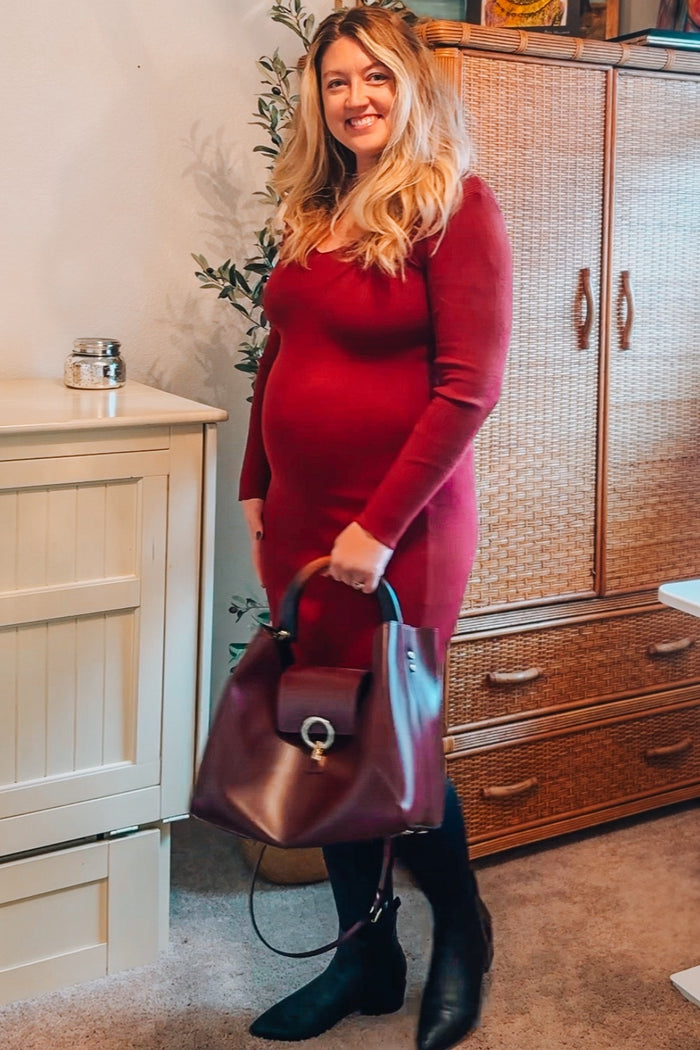 woman wearing a burgundy long sleeve maternity dress, holding a burgundy handbag and showing her baby bump