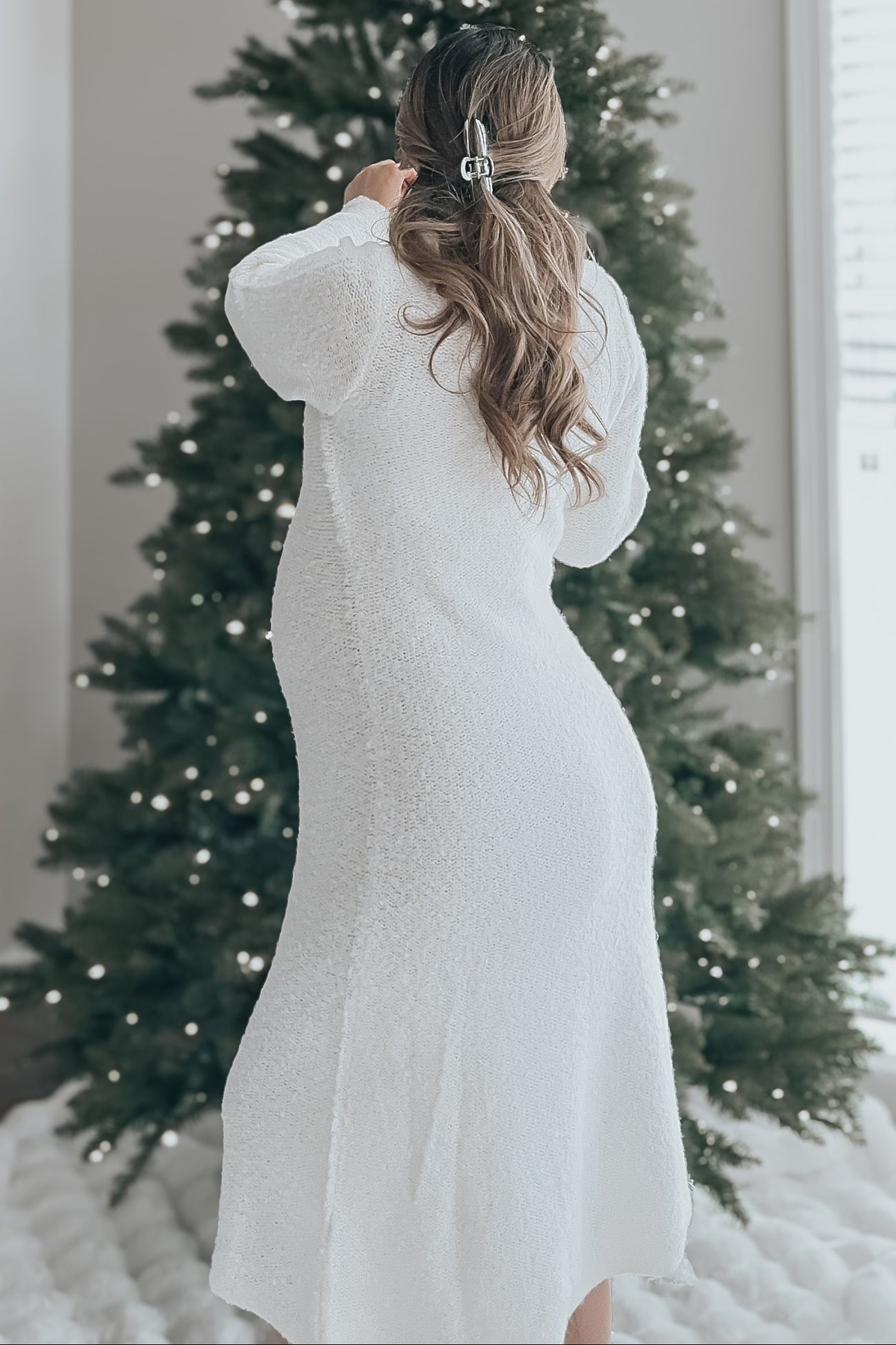 woman wearing a white maternity dress with long sleeves, standing with a baby bump in front of a decorated Christmas tree