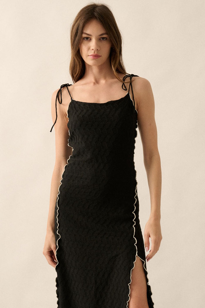 woman wearing a black maxi dress with a textured pattern and white trim detailing, featuring adjustable straps and a side slit