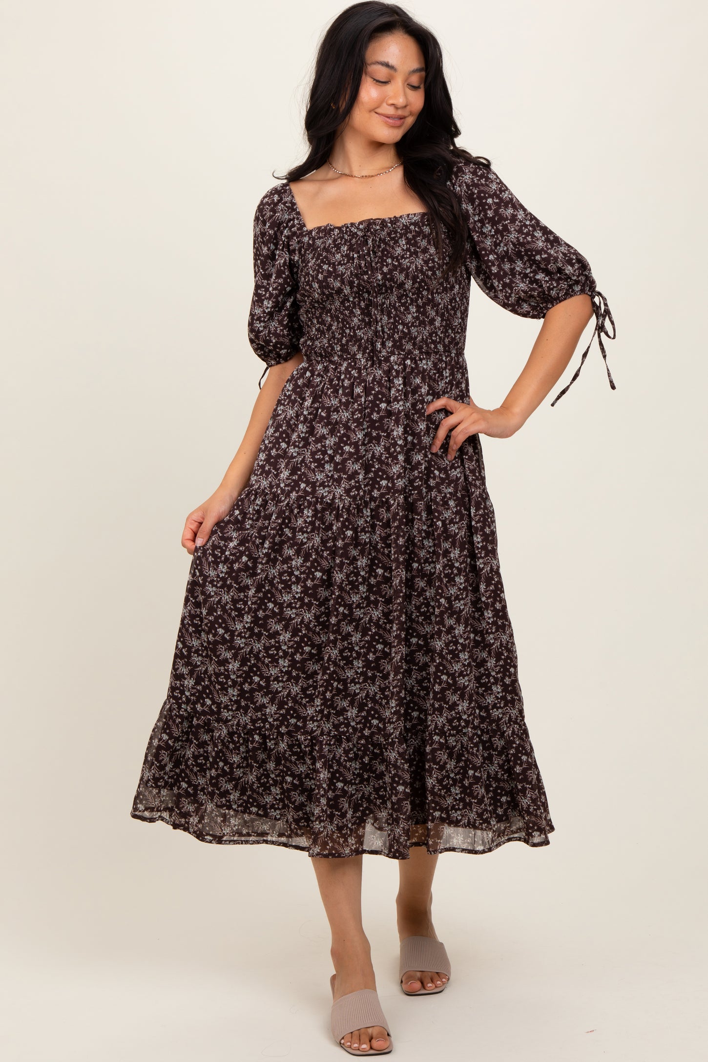 woman wearing a dark floral midi dress with puff sleeves and tie details on the sleeves, standing on a neutral background