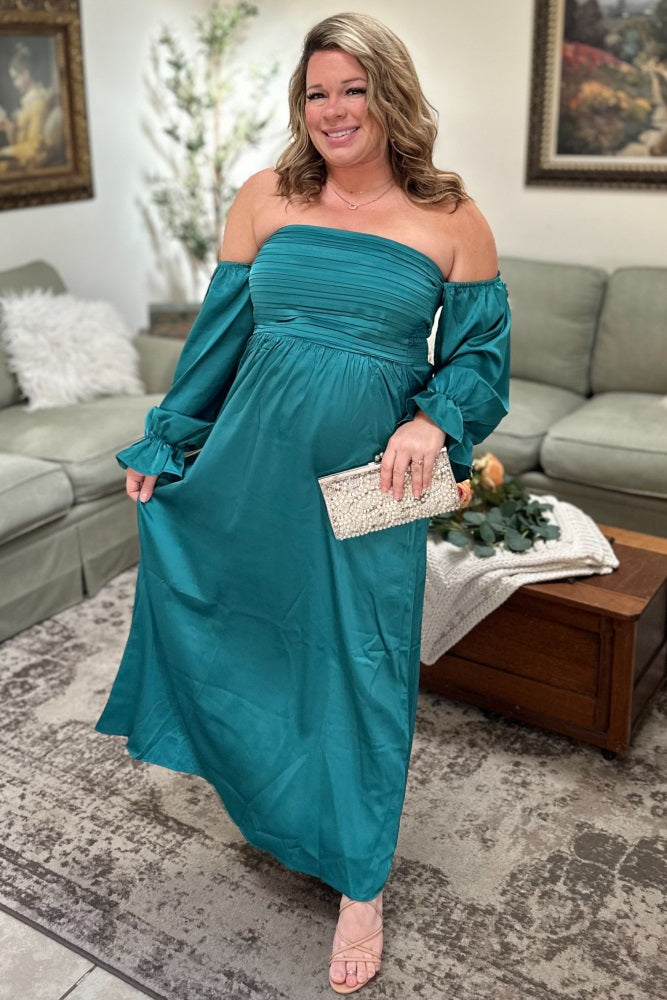 woman wearing a teal off-the-shoulder maxi dress with gathered bodice and long sleeves, standing in a living room