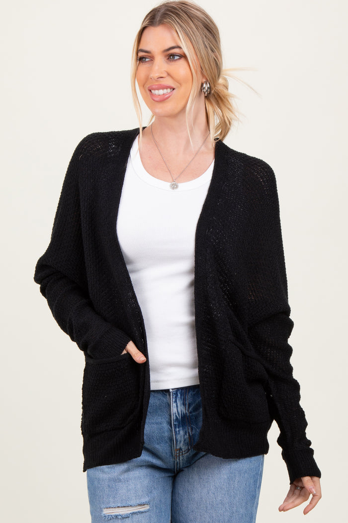 woman wearing a black open-knit cardigan over a white tank top and blue jeans, smiling with hands in pockets
