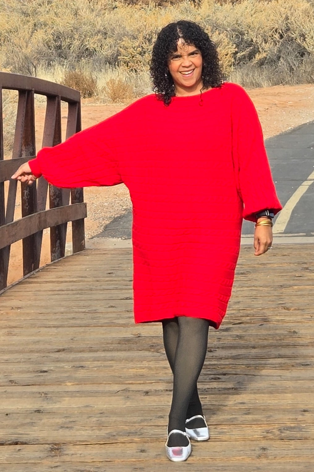 woman wearing a bright red oversized sweater dress with three-quarter sleeves and black tights, walking on a path