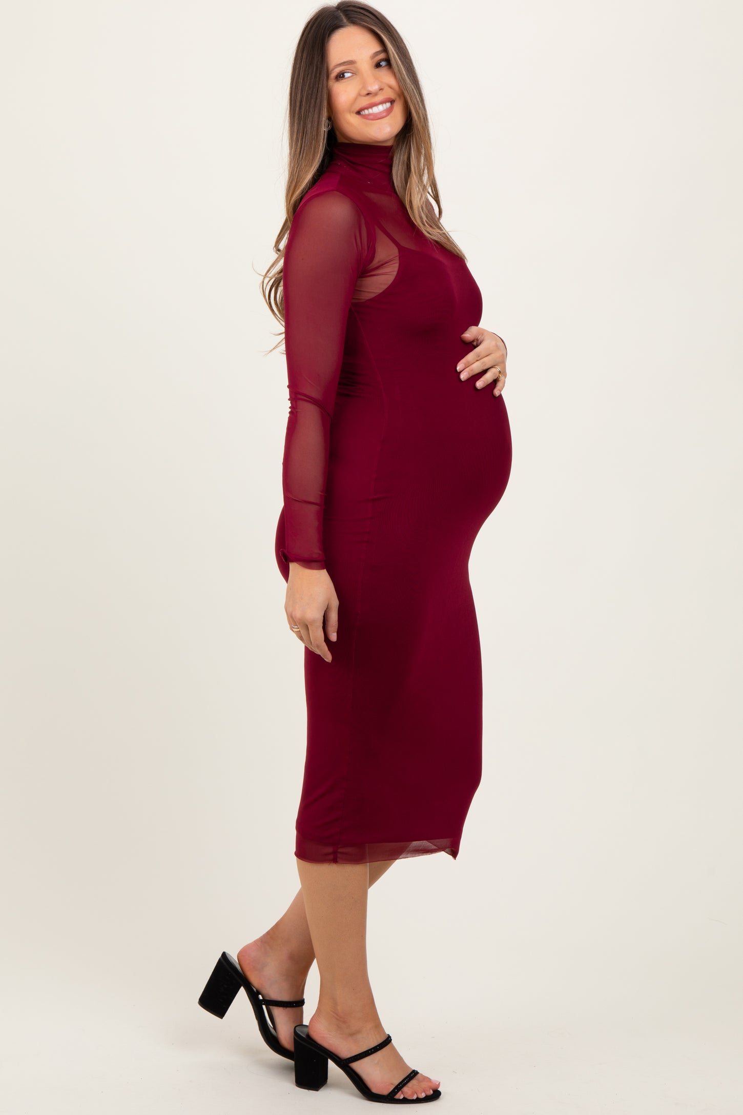 woman wearing a burgundy long sleeve maternity dress, highlighting her baby bump and styled with strappy black heels