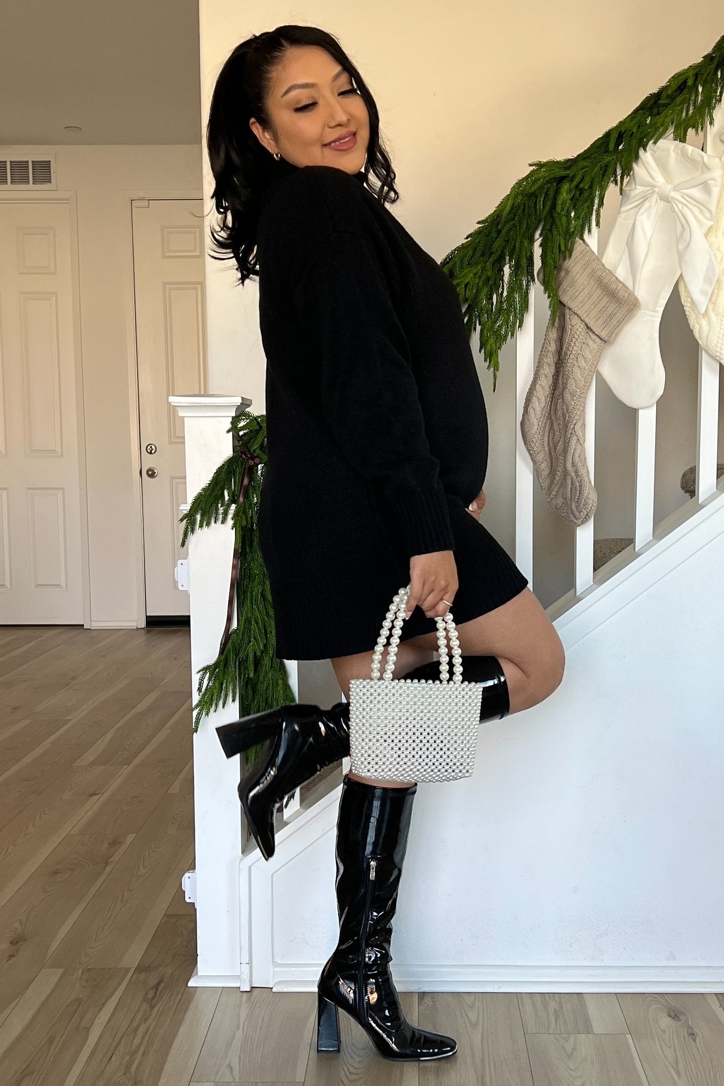 woman wearing a black oversized sweater dress with knee-high black boots and holding a small pearl handbag, showing her baby bump
