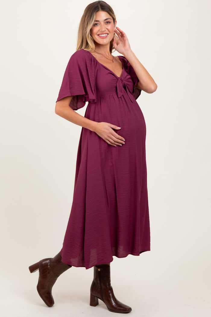 woman wearing a burgundy maternity dress with a tie front and flutter sleeves, gently cradling her baby bump