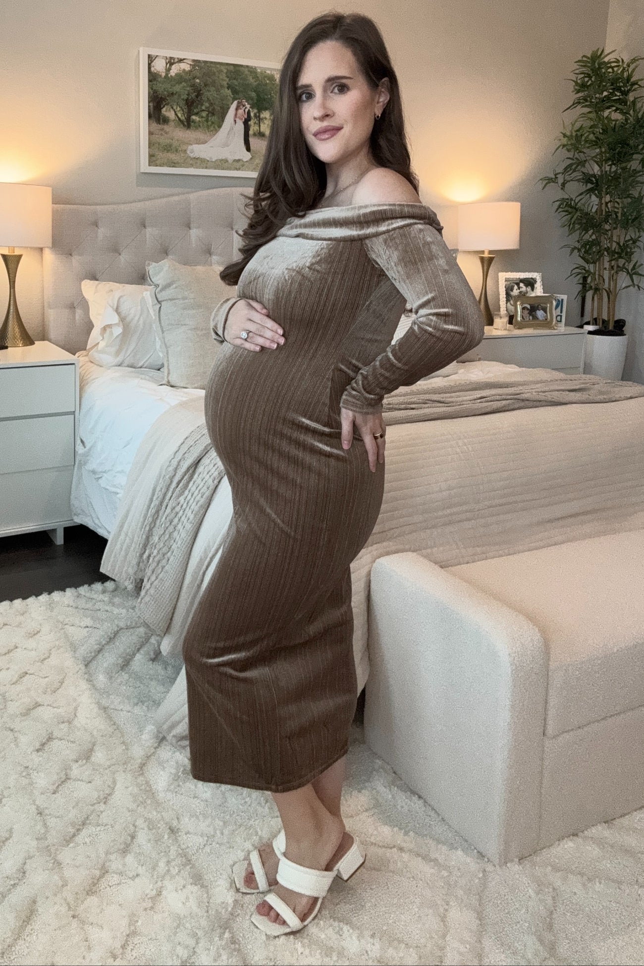 woman wearing a brown off-shoulder maternity dress with a visible baby bump, standing in a bedroom setting