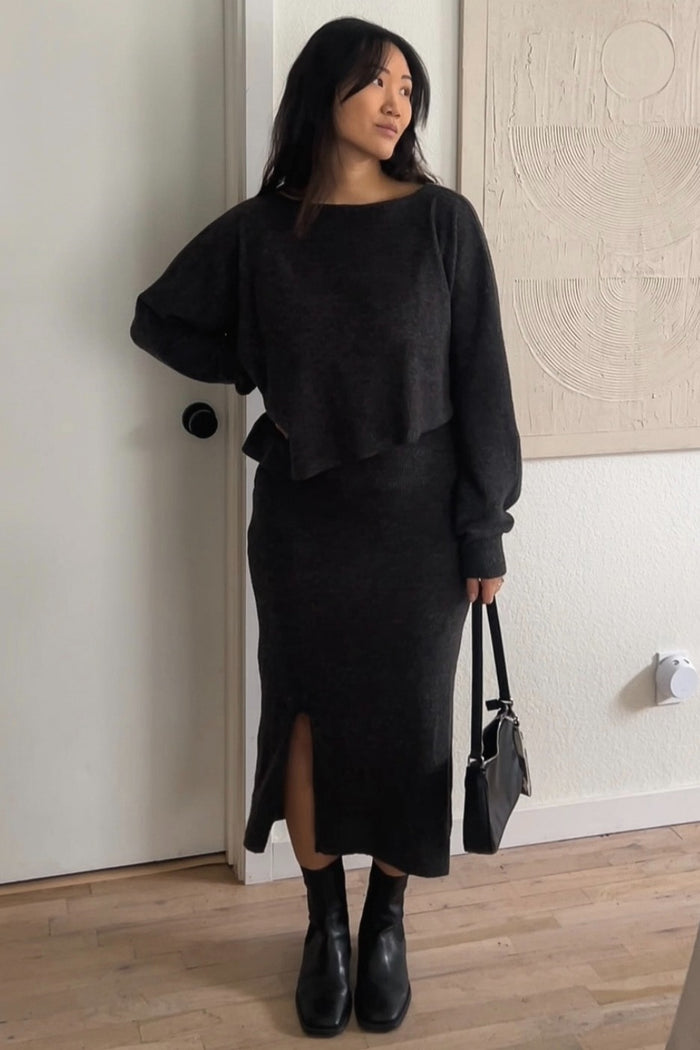 woman wearing a dark gray cropped sweater and matching long skirt with a slit, holding a small black handbag