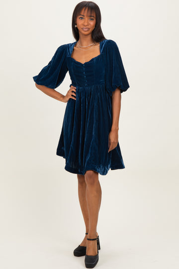 woman wearing a navy blue velvet short sleeve dress with a square neckline and button details, standing in black heels