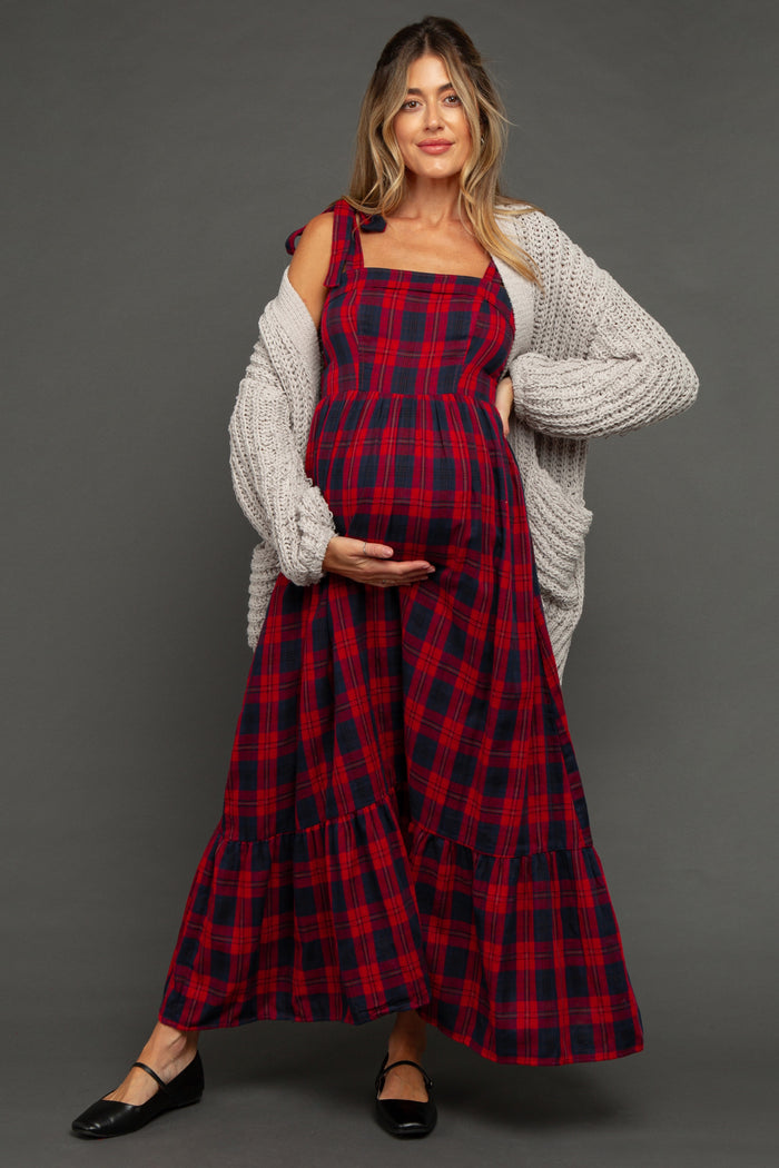 woman wearing a red and navy plaid maternity dress with a ruffled hem and a cozy gray cardigan, holding her baby bump