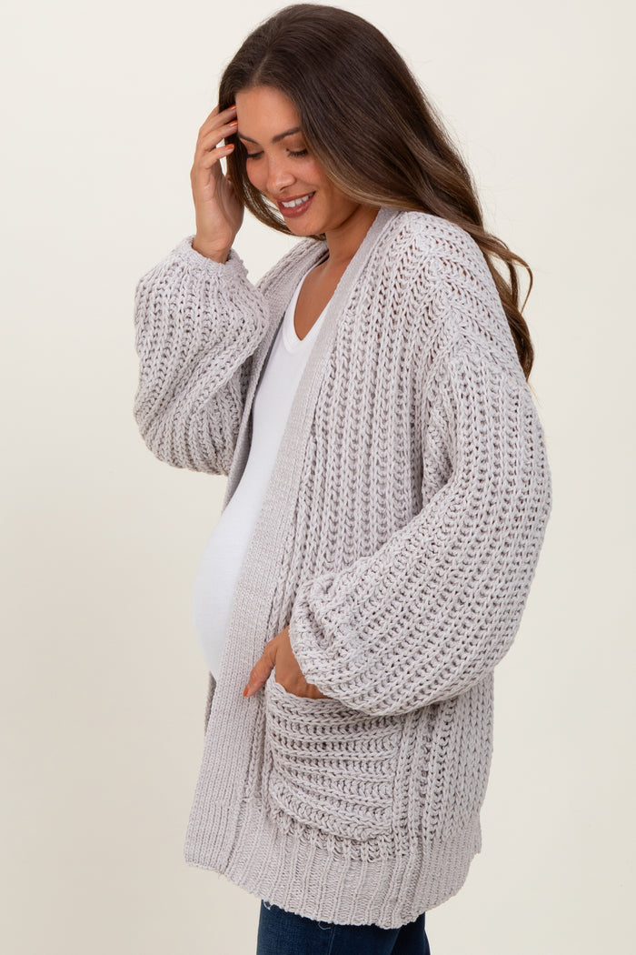 woman wearing a light grey knitted maternity cardigan with pockets, gently touching her hair and showing her baby bump