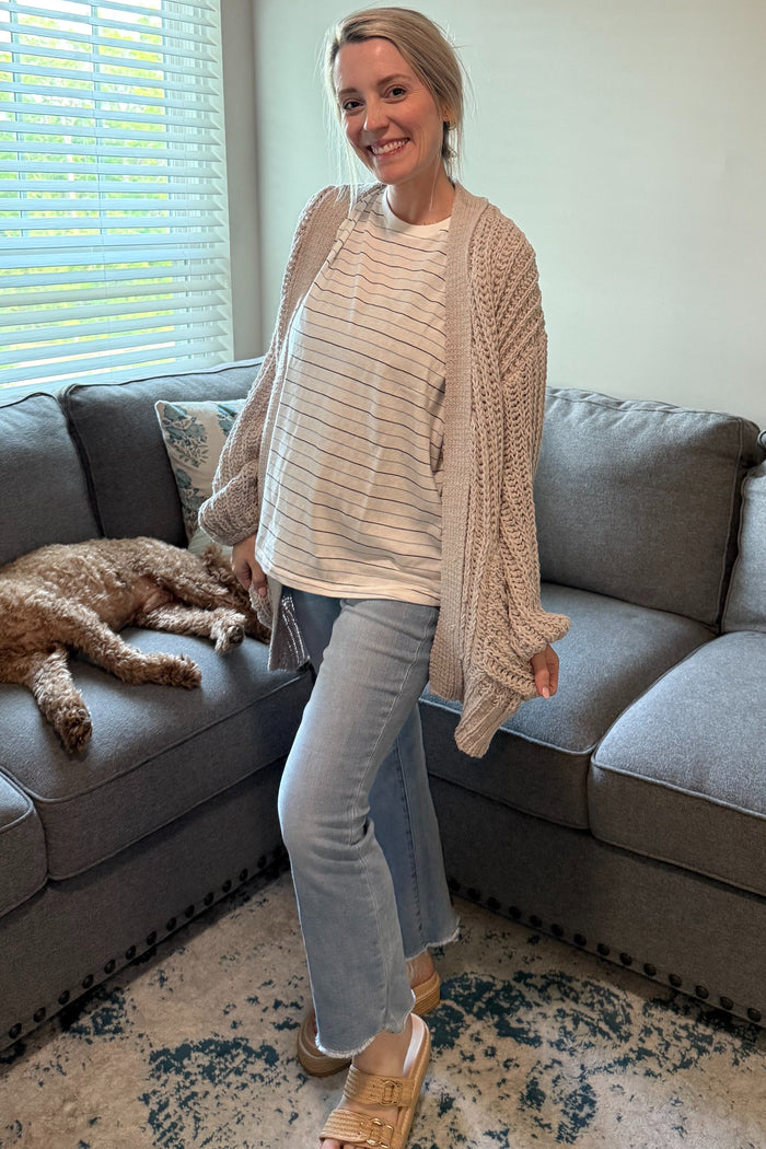 woman wearing a cream striped maternity top and a chunky beige cardigan, paired with light jeans and sandals, standing on a sofa