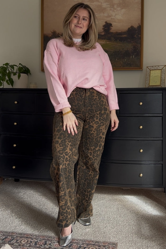 woman wearing a pink sweater and leopard print pants, standing in front of a dresser and smiling