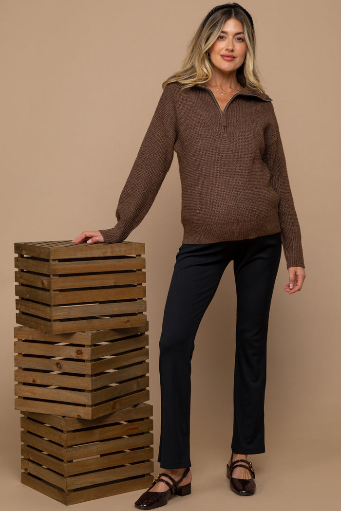 woman wearing a dark brown zip-up maternity sweater and black maternity pants, posing with wooden crates