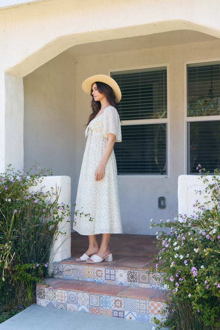 woman wearing a floral midi dress with a tie front, straw hat, and wedge sandals, standing on a porch surrounded by plants