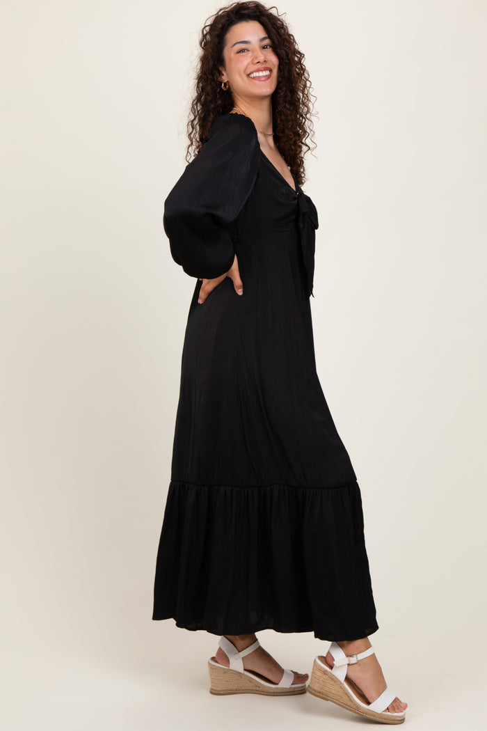 woman wearing a black maxi dress with puff sleeves and a tied neckline, paired with white wedge sandals