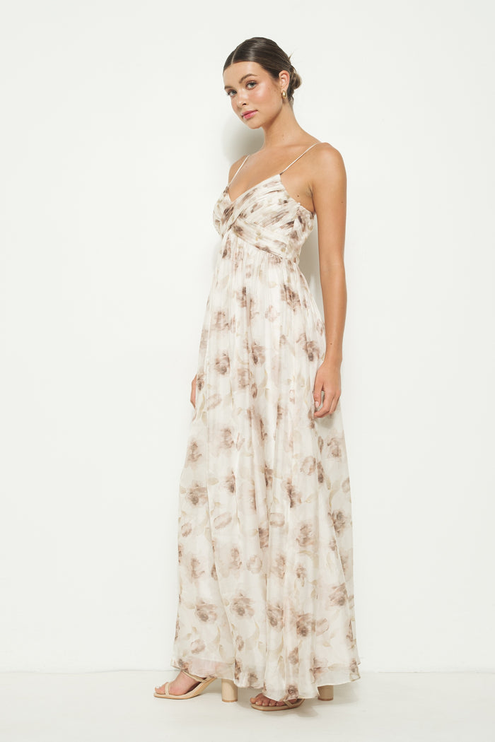 woman wearing a sleeveless maxi dress with a floral pattern in beige and brown tones, standing against a white background