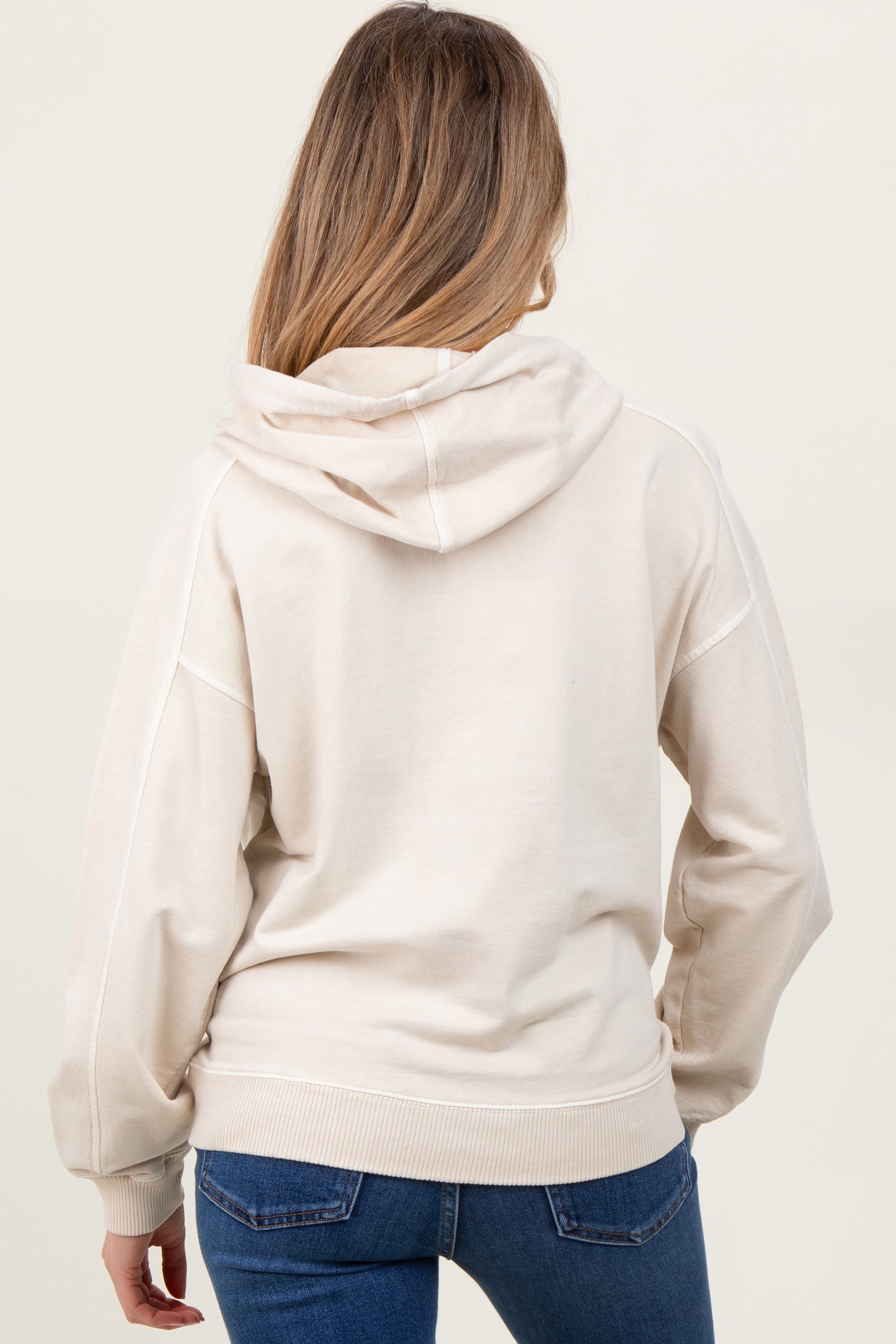 woman wearing a light beige maternity hoodie with a relaxed fit and a hood, showing her back and styled with jeans