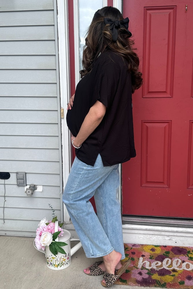 woman wearing a black maternity top and light blue wide-leg jeans, showing her baby bump while standing by a door