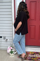 woman wearing a black maternity top and light blue wide-leg jeans, showing her baby bump while standing by a door
