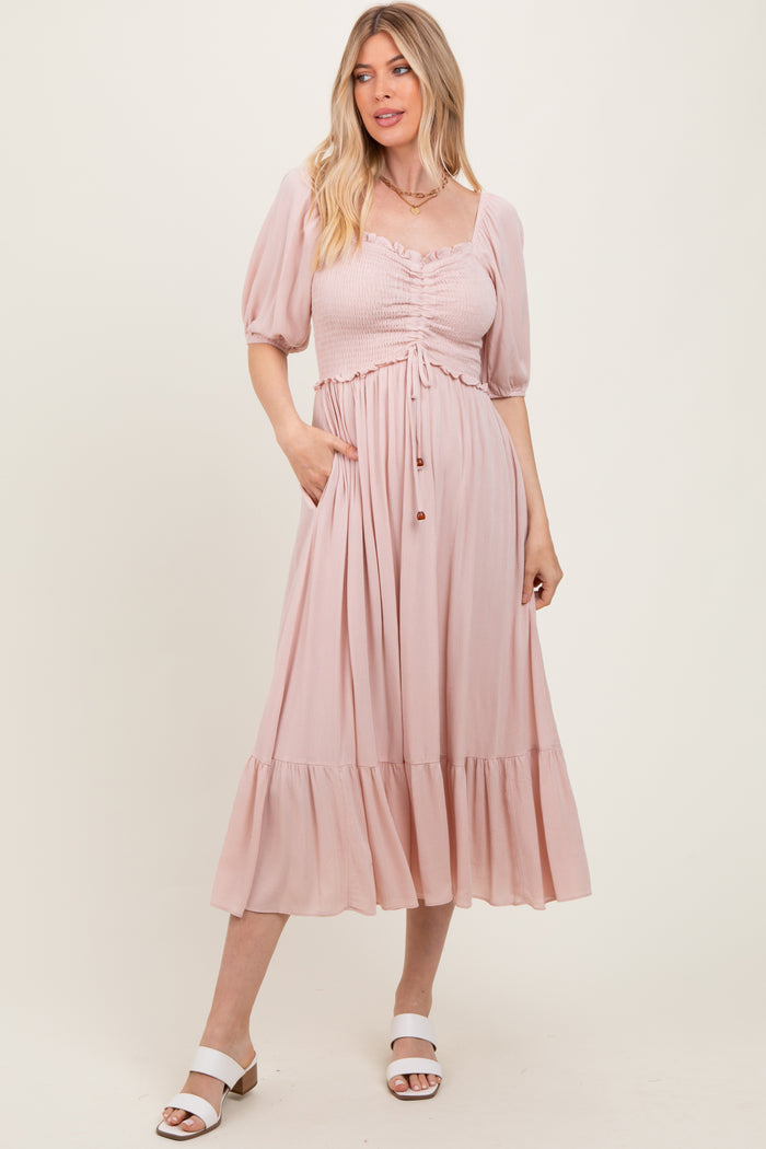 woman wearing a light pink midi dress with puff sleeves, a smocked bodice, and a tiered hem, standing and smiling