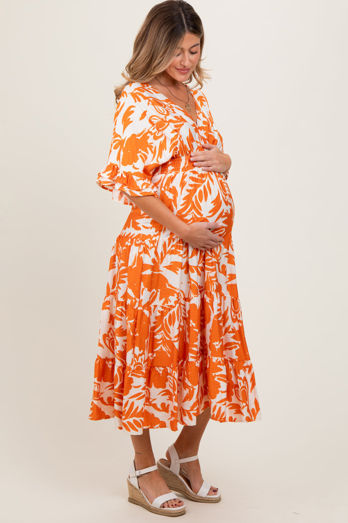woman wearing an orange and white floral maternity dress and cradling her baby bump